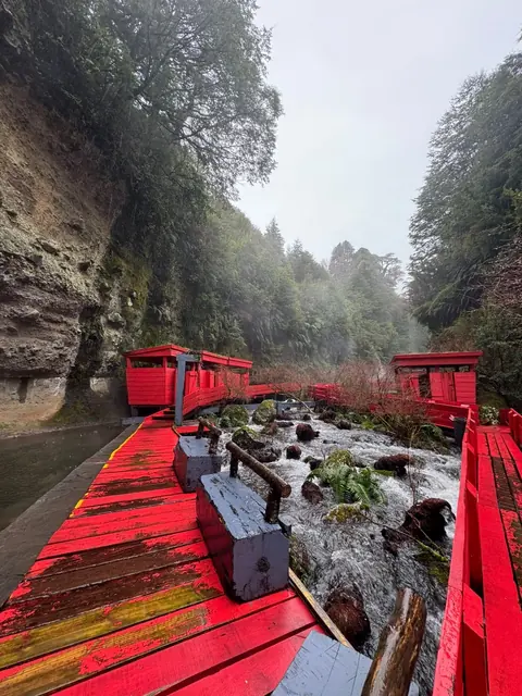 Pasarela roja al lado de un curso de agua con rocas y construcciones de madera