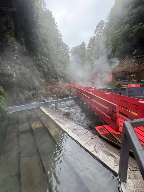 Pasarela con barandas grises y rojas sobre agua y rocas en la quebrada